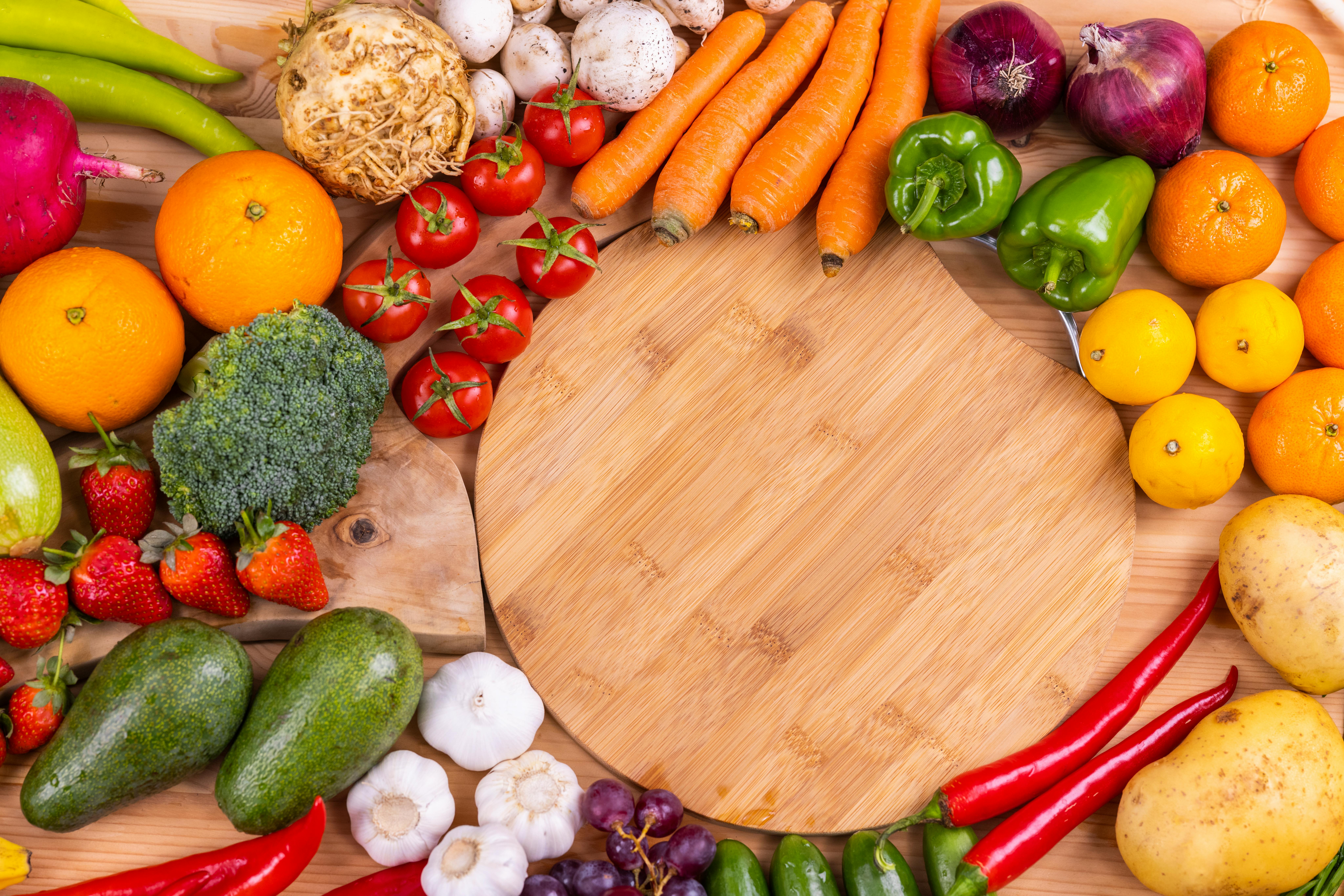 Fresh fruits and vegetables surrounding a wooden cutting board, including carrots, broccoli, tomatoes, avocados, oranges, peppers, garlic, and strawberries on a wooden table.